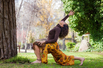 Mature woman practices yoga early morning in the park on sunny day. Wearing colorful pants and brown long sleeve shirt on warrior pose, known as virabhadrasana