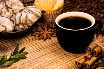 On a wooden table a mug of black coffee next to a plate of chocolate cookies sprinkled with powdered sugar, and on the background coffee beans spread with cinnamon, puddings and green coffee twing