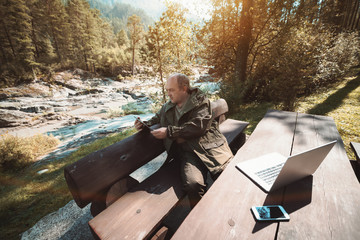 Wide-angle photo of mature forest-guard sitting on wooden bench at the table and having online...