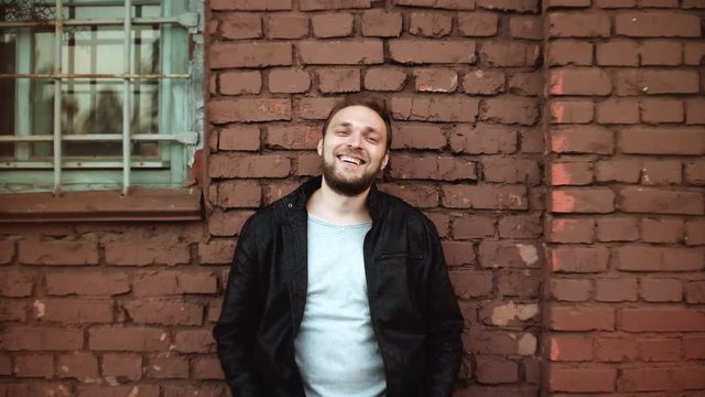 Excited handsome European man smiles into camera. Happy successful male businessman stands near red brick wall 4K.