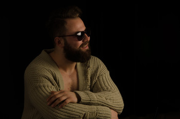 The young model posing in the studio is dressed in a cardigan and has bearded in his pockets and bearded .. sitting on a chair, looking forward and happy
