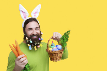 Happy Easter concept. Nice kind muscle man male with beard, white ears of rabbit, carrots and basket with colorful easter eggs and green smiling in green t-shirt isolated on yellow background