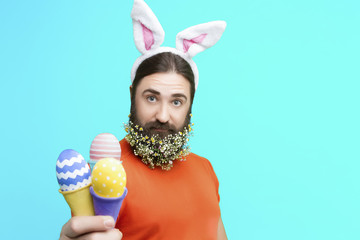 Happy Easter concept. Nice kind muscle man male with flower's beard  with white ears of rabbit and colorful easter eggs in waffle in orange t-shirt isolated on blue background