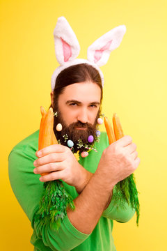 Happy Easter concept. Nice kind muscle man male with spring's flower's beard, white ears of rabbit, carrots in green t-shirt isolated on yellow background