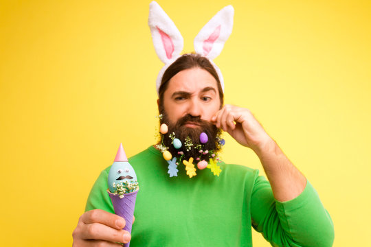 Happy Easter concept. Nice kind muscle man male with beard pined by easter eggs, white ears of rabbit and comic blue egg in waffle in green t-shirt isolated on yellow background