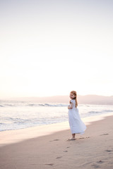 Young girl in white dress standing on the beach looking back at the camera from a distance