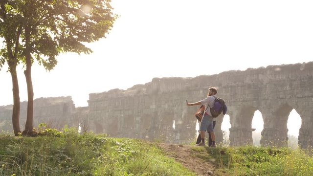 Young lovely couple backpackers tourists taking selfies photo on hill in front of roman aqueduct arches in parco degli acquedotti park ruins in rome on romantic misty sunrise with sleeping bag slow