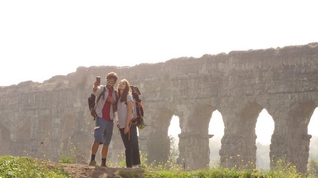 Young lovely couple backpackers tourists taking selfies photo on hill in front of roman aqueduct arches in parco degli acquedotti park ruins in rome on romantic misty sunrise with sleeping bag slow