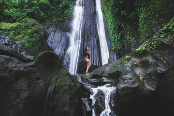 Woman enjoying near hidden in jungle cascade waterfall in Bali. Slim body and black swimsuit, fashion model. Dusun Kuning in Ubud area