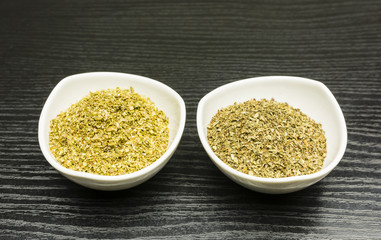 White bowls with dried basil and oregano on a dark table.