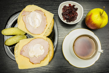 Plate with a slice of bread with ham, cheese and mayonnaise, for this cucumber, and an apple and a bowl of dried cranberries. Drinking a cup of tea.. View from above.
