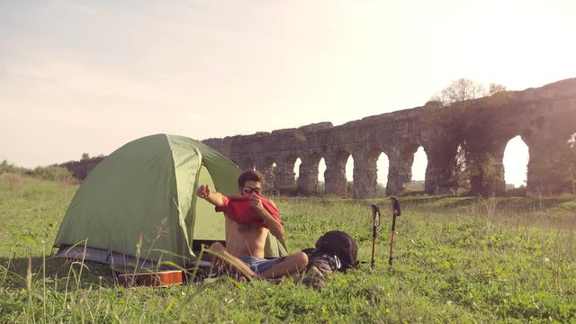 Young man backpacker dressing wearing t-shirt outside camping tent sitting on grass in front of roman aqueduct arches in parco degli acquedotti park ruins in rome at sunset sticks in ground slow
