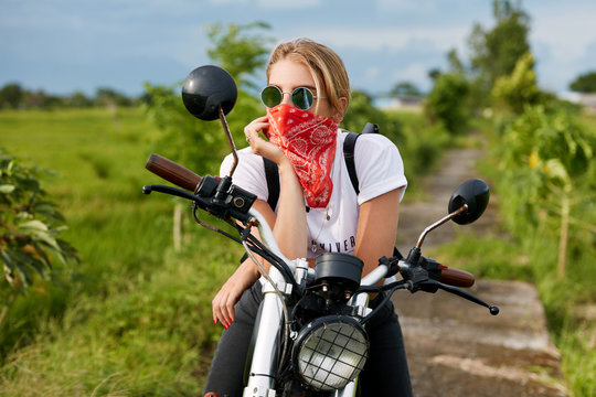 Attractive female in casual clothing, sits on motorbike, enjoys freedom and active lifestyle, has thoughtful expression, poses against green plantation backround outdoor. Motorcycle tourism concept