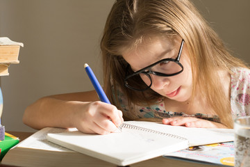 A little girl in glasses is sitting at a table and writing in a notebook