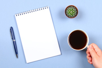 Empty notebook with pen, coffee and cactus on a blue background. Copy the space. Top view. A hand holds a cup of coffee.