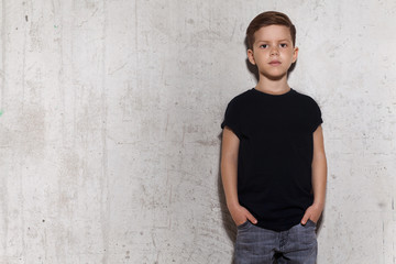 Cute little boy in black T-shirt posing in front of grunge concrete wall. Portrait of fashionable male child, copy space. Boy looks at camera, gray wall on background. © deniskomarov