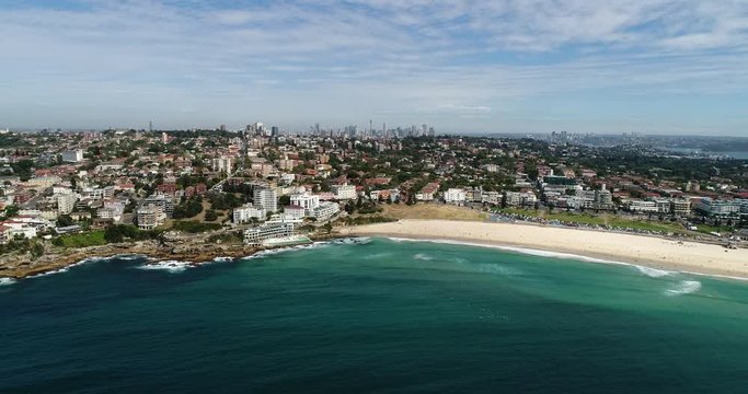 Bondi beach waterfront and sand in backward aerial flying with distant Sydney city CBD on horizon.
