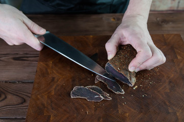 Portion of Beef Jerky cutting into slice on a cutting table with dark beer glass and a kitchen knife on vintage wooden background, man standing behind