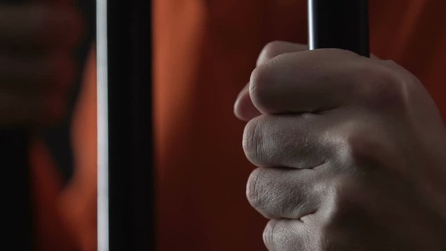 Angry prisoner in orange jumpsuit holding cell bars, serial killer hands closeup
