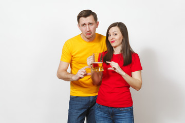 Sad upset crying young couple, woman, man, football fans in yellow red t-shirts with pint mug of little beer, show fingers ends isolated on white background. Sport, family leisure, lifestyle concept.