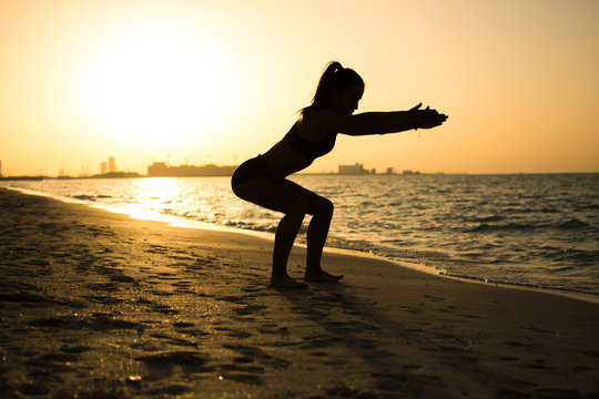Silhouette of slim sporty girl exercising on the beach during sunset.