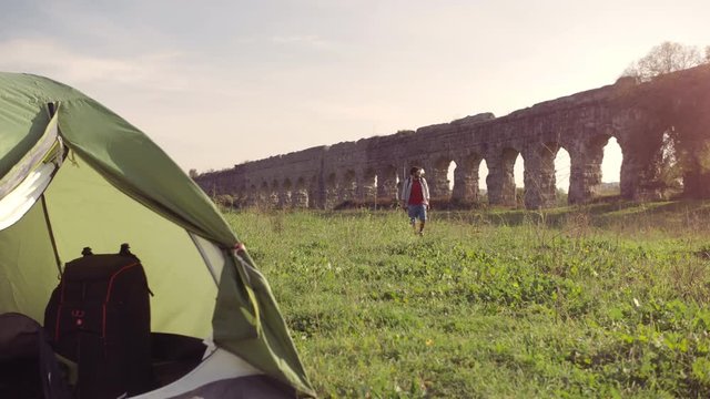 Young man backpacker outside camping tent walks barefeet with sticks on grass in front of roman aqueduct arches in parco degli acquedotti park ruins in rome at sunset slow motion