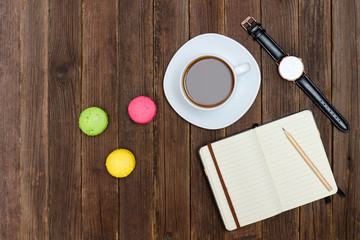 Top view on cup of coffee, macarons, notebook and watch. Wooden background