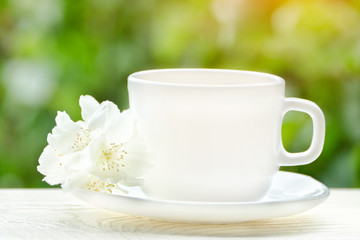 White mug of tea with jasmine on a background greenery, sunlight. Close up