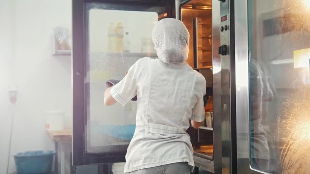 Female bakes on commercial kitchen - pulls the bread from the oven