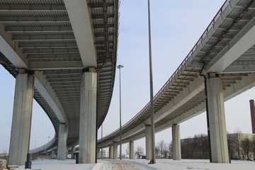 The huge concrete bridge for cars on huge support standing on the earth covered snogy against the background of the gray winter sky in a hard frost.