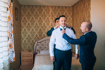 stylish groomsmen helping happy groom getting ready in the morning for wedding ceremony. luxury man...