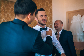 stylish groomsmen helping happy groom getting ready in the morning for wedding ceremony. luxury man in suit in room. space for text. wedding day.