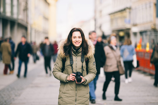 portrait Beautiful young tourist woman stands in the background of a crowd of people on a central street in Munich in Germany in winter. Holds a black big professional camera and smiles.