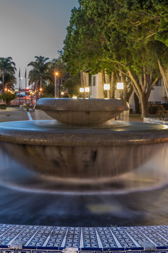 Street lamps shining over water fountain as dawn light begins to illuminate the sky.