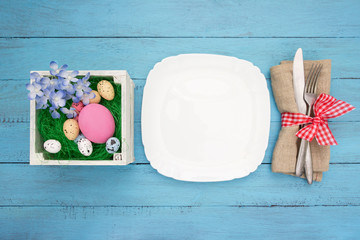White plate,fork and knife at napkin,easter nest with eggs  on blue wooden board.Easter table set with space for text