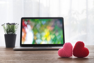Office table with Beautiful couple knitted fabric heart shape, heart shape bokeh background on laptop screen and violet flower pot. view from front office table. valentine's day concept.