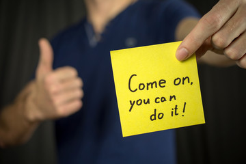 Man holds yellow sticker inscription Come on, you can do it! Thumb up Blue T-shirt. close-up, selective focus