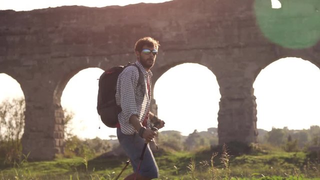Young man backpacker walks barefeet with sticks on grass in front of roman aqueduct arches in parco degli acquedotti park ruins in rome at sunset slow motion