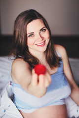 Attractive young pregnant woman sits on a bed and eats a ripe strawberry. Food for a happy pregnant girl, fruits and vegetables for a balanced diet future mom. Healthy food for a successful pregnancy.