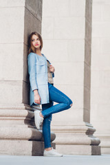 the girl stands with a raised leg, in a blue coat and jeans and white sneakers