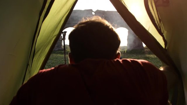 Young man backpacker exits camping tent in front of roman aqueduct arches in parco degli acquedotti park ruins in rome at sunset sticks in ground shot from inside tent camera slow motion pov
