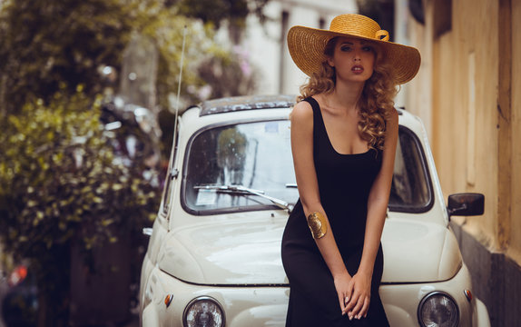 Elegant portrait of beautiful fashion woman wavy shine blonde long hair model in summer hat black dress and luxury accessories with bright makeup beside retro car in Trastevere Roma Italy. Soft light