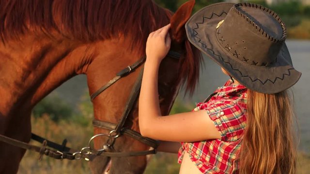 Portrait of a young girl next to the horse on the background of a small lake and forest in summer at sunset.