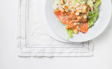 Leaf vegetable salad with smoked salmon, parmesan cheese and bread crumbs in a white plate on a light wooden background. View with copy space. Flat lay, top view