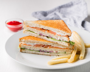 Freshly made club sandwiches with French fries served on white plate. Pressed double toasts with leaf vegetable salad, smoked salmon and cheese served on paper on a wooden table. Street fast food.