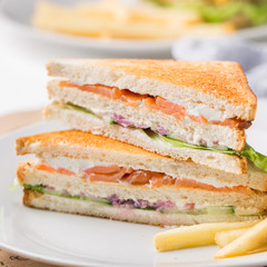 Freshly made club sandwiches with French fries served on white plate. Pressed double toasts with leaf vegetable salad, smoked salmon and cheese served on paper on a wooden table. Street fast food.