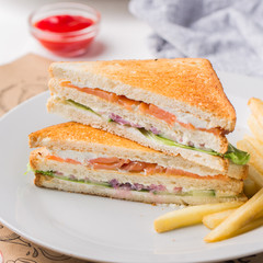 Freshly made club sandwiches with French fries served on white plate. Pressed double toasts with leaf vegetable salad, smoked salmon and cheese served on paper on a wooden table. Street fast food.