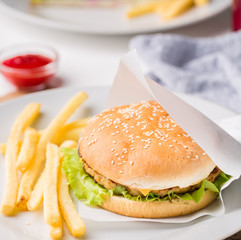 Fresh tasty burger and french fries served on white plate. Delicious burger with beef, tomato, cheese and lettuce served on paper on a wooden table. Street fast food