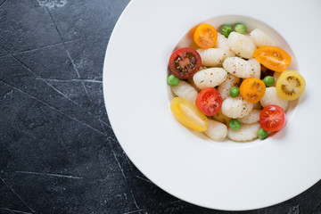 White plate with italian gnocchi, fresh tomatoes and green peas, view from above on a grey concrete background, horizontal shot with space