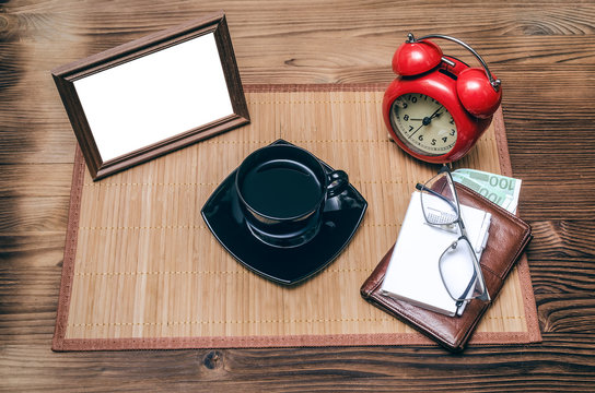 Photo frame with copy space, red alarm clock and cup of black coffee and wallet with money on office desk table background. Coffee break.